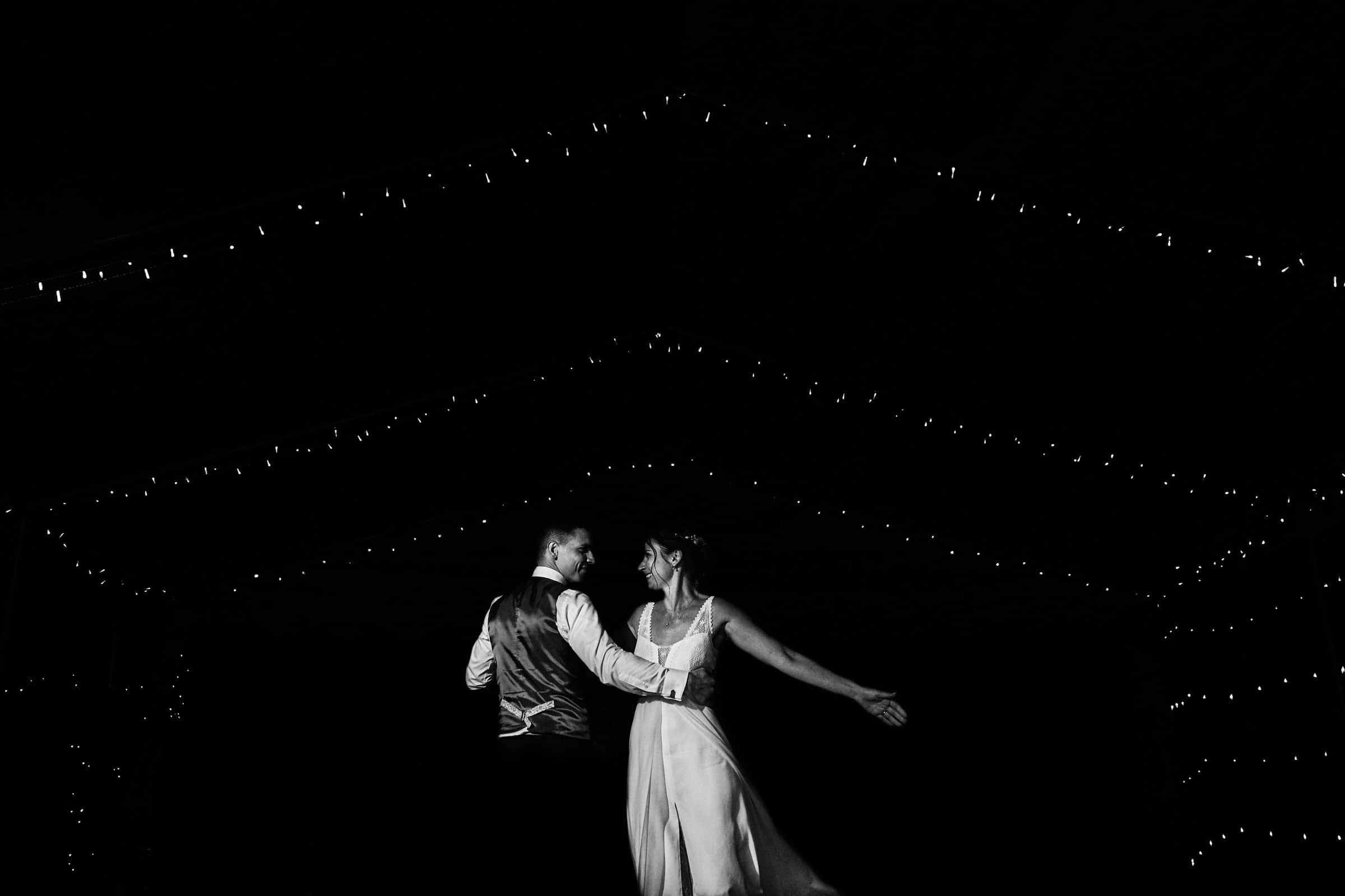Mariés dansant sous des guirlandes lumineuses, en noir et blanc